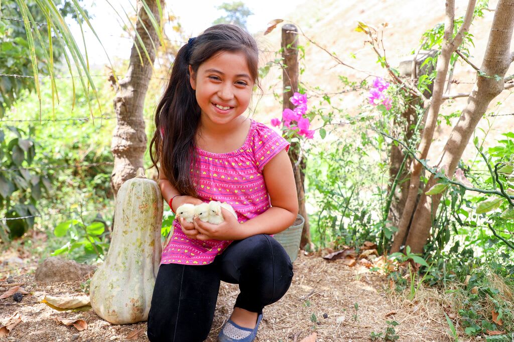 Little girl with a pink shirt holds 3 baby chicks in her hands smiling.